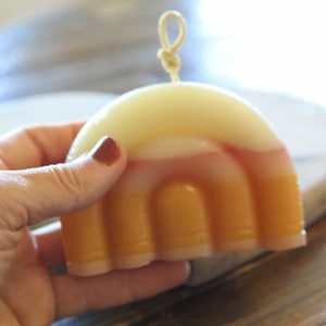 Könnte beinhalten: Eine mehrfarbige Kerze mit einem mehrschichtigen Design in Weiß, Rosa und Orange. Die Kerze ist bogenförmig und hat oben einen Docht.