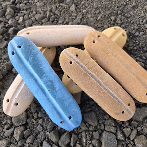 May include: A collection of elongated, smooth stones in various colors, including blue, beige, and tan. Each stone has a central groove and two drilled holes. The stones are arranged on a bed of small, gray rocks.