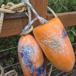 May include: Two orange buoys with blue and white paint splotches hang from a wooden fence post. The buoys are attached with rope and show signs of wear. The background features green foliage and additional rope.