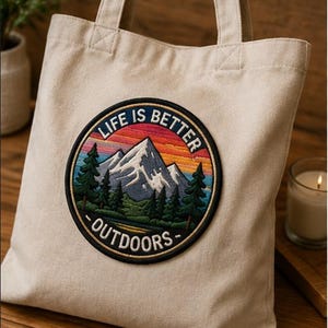 May include: A cream-colored canvas tote bag featuring a circular embroidered patch. The patch depicts a mountain landscape with green pine trees, a lake, and a sunset sky in shades of orange and blue, with the text LIFE IS BETTER OUTDOORS.