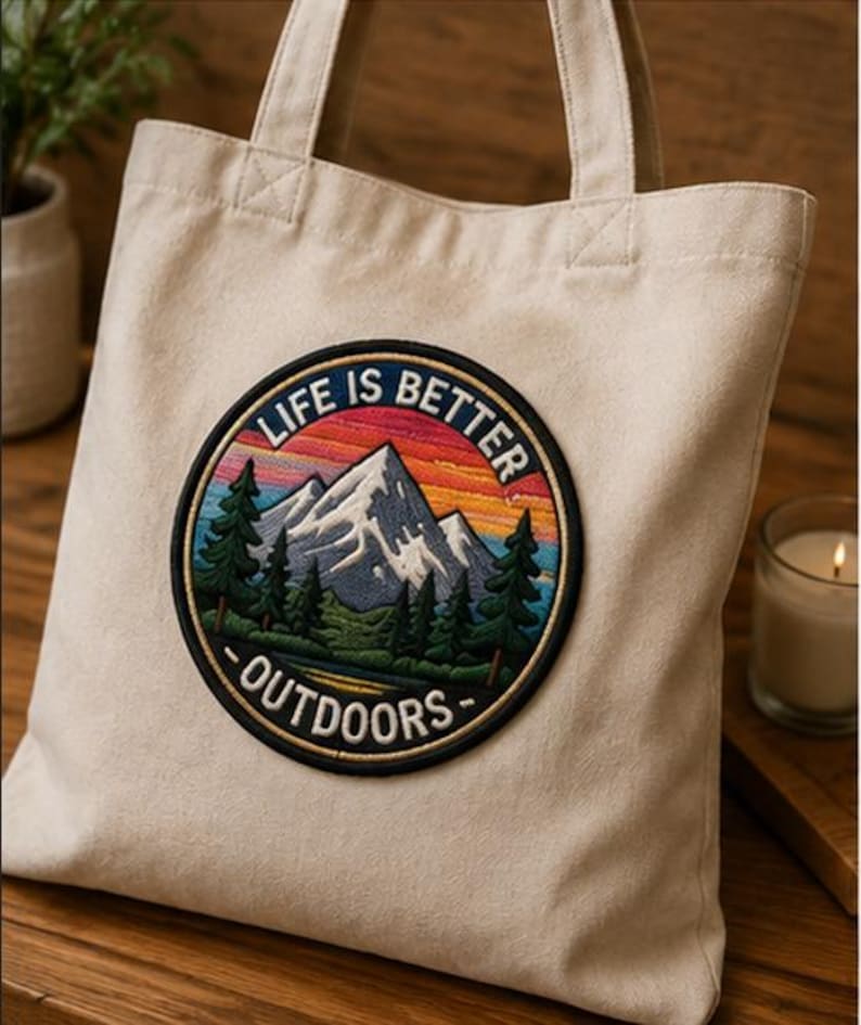 May include: A cream-colored canvas tote bag featuring a circular embroidered patch. The patch depicts a mountain landscape with green pine trees, a lake, and a sunset sky in shades of orange and blue, with the text LIFE IS BETTER OUTDOORS.