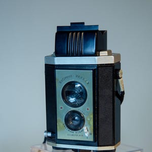 May include: A vintage Brownie Reflex camera, black and silver, with the text "Brownie Reflex" and "Eastman Kodak Rochester, N.Y." on the front. A booklet titled "How to use the Brownie Reflex" is in front of the camera.