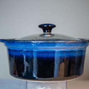May include: A ceramic casserole dish with a lid. The dish has a gradient of blue hues, transitioning from a lighter shade at the top to a deep navy at the base. The lid has a black knob.