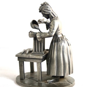 Könnte beinhalten: Eine Zinnfigur einer Frau, die Flüssigkeit aus einer Schöpfkelle in eine Form gießt. Die Frau trägt ein langes Kleid und eine Mütze. Die Form steht auf einem Tisch mit einem Sockel. Die Figur steht auf einem runden Sockel.