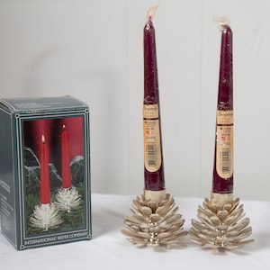 May include: Two burgundy taper candles in silver pinecone-shaped holders. A box with a similar design is on the left. The box features a picture of the candles and holders, with the text "INTERNATIONAL SILVER COMPANY".