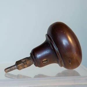 May include: A dark brown, rounded door knob with a smooth, polished finish. The knob features a metallic, threaded spindle extending from the base, which includes a small, visible screw for secure attachment to a door mechanism.