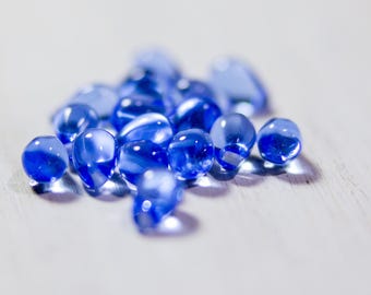 May include: A group of small, round, blue glass beads on a white surface.