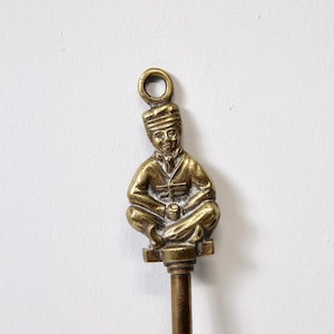 May include: A vintage brass key with a decorative seated figure on top. The figure is wearing a hat and appears to be holding an object. The key has a loop at the top and a long, slender stem.