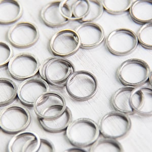 May include: A collection of small, silver-colored metal rings scattered on a white surface. The rings are circular with a hollow center, and appear to be made of a matte metal. The lighting creates subtle shadows, highlighting the texture of the rings.