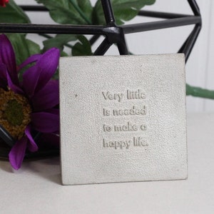 May include: A white square concrete paperweight with the text "Very little is needed to make a happy life." embossed on the surface.