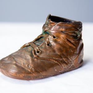May include: A bronze-colored sculpture of a baby's high-top shoe. The shoe has laces and a textured surface, with hints of green patina. The sculpture is set against a white background.