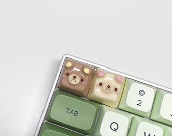 May include: A close-up of a mechanical keyboard with custom keycaps. Two keycaps feature bear designs, one brown and one cream-colored. The keyboard has green and white keys.
