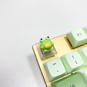 May include: A green frog keycap with white eyes and a yellow belly sits on a light green keyboard key. The keyboard is gold and has a light green F1 key.