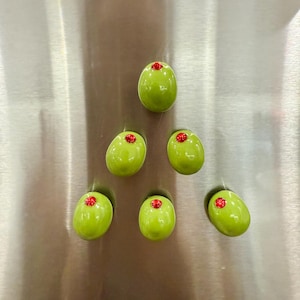 May include: Six green olive-shaped magnets with red accents are displayed on a silver surface. The magnets have a glossy finish and appear to be ceramic or resin. The red accents are small and circular, resembling pimentos.