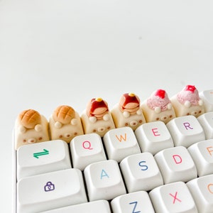 May include: A white keyboard with colorful keycaps featuring miniature food designs. The keycaps are shaped like a pancake, a donut, a strawberry, a cupcake, and a croissant. The keyboard has keys labeled QWERTYASDFGHJKLZXCVBNM.
