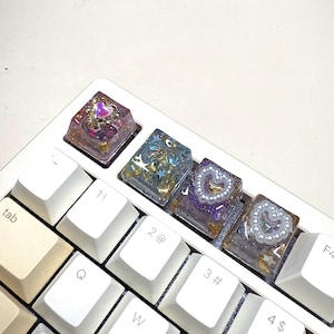 May include: Close-up of a white keyboard with custom resin keycaps. The keycaps are clear with embedded designs, including hearts, stars, and gold flakes. The keyboard keys are white and off-white.
