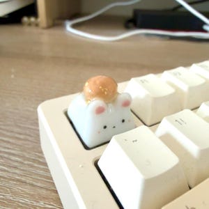 May include: A white custom mechanical keyboard keycap shaped like a small animal with pink ears, black eyes, and a light brown circular accent on top. The keycap is installed on a white keyboard among other standard white keys on a wooden surface.