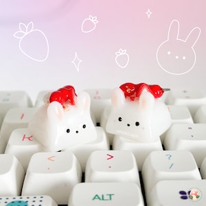 Peut inclure: Deux touches de clavier blanches en forme de lapin avec des accents de fraises rouges et des détails de feuilles d'or. Les touches sont sur un clavier blanc. Le fond présente des fraises et un visage de lapin dessinés en blanc.