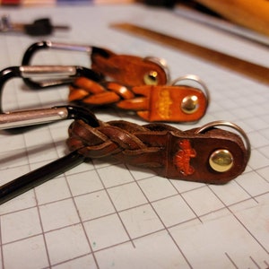 May include: Three leather keychains with braided leather straps and metal key rings. The keychains are brown and have a gold-colored metal button.