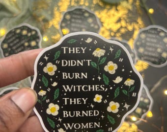 Può includere: Un adesivo nero con testo bianco che recita: "THEY DIDN'T BURN WITCHES, THEY BURNED WOMEN." L'adesivo ha un design floreale con fiori bianchi e foglie verdi.
