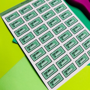 May include: A sheet of stickers with a green and white design. Each sticker has the words "PAY DAY" and a dollar sign. The stickers are rectangular with a light green border. The background is a mix of green and yellow.