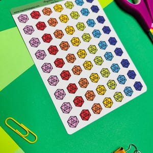 May include: A sheet of colorful D20 dice stickers in various colors, including red, orange, yellow, green, blue, and purple. Each sticker features a 20-sided die design with the number 20. The sheet is on a green surface with office supplies.