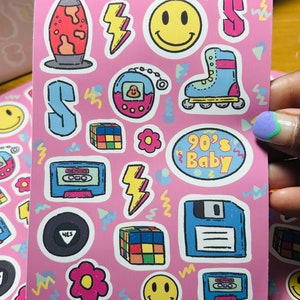 Puede incluir: Una hoja de pegatinas rosas con temática de los 90. Las pegatinas presentan una variedad de imágenes, incluyendo una lámpara de lava, una cara sonriente, una cinta de casete, un disquete, un cubo de Rubik, una flor, un rayo, un Tamagotchi, un par de patines y el texto "90's Baby".