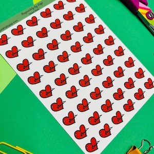 May include: A sheet of stickers featuring red heart illustrations with a heartbeat line. The stickers are on a white background, with the text "Roquishly by Gigi Heart Rate" visible. A pair of purple scissors and a yellow paper clip are also in the image.