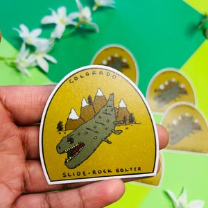Può includere: Un adesivo con un'illustrazione a cartoni animati di una creatura rettiliana verde, con l'etichetta "SLIDE-ROCK BOLTER" e la parola "COLORADO" sopra. L'adesivo ha uno sfondo giallo con montagne e alberi bianchi.