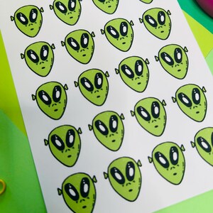May include: A white sticker sheet featuring twenty-one identical green alien head illustrations with large black eyes and small noses. The text "Roguishly by Gigi" and "Alien" is printed at the top, along with the website "www.roguishlybygigi.com".