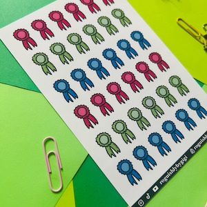 May include: A sheet of stickers with 36 colorful ribbon award designs. The stickers are in three colors: blue, green, and pink. The stickers are arranged in rows on a white background.