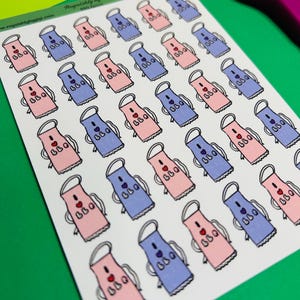 Könnte beinhalten: Ein Bogen Aufkleber mit Schürzenmotiven in Rosa und Blau mit dem Text "BBQ" und einem Herz. Die Aufkleber haben einen weißen Hintergrund und sind in Reihen und Spalten angeordnet. Der Bogen liegt auf einer grünen Oberfläche.