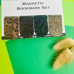 Może przedstawiać: Zestaw czterech magnetycznych zakładek z wzorem. Zakładki są brązowe, czarne i zielone z złotym wzorem. Tekst "MAGNETIC BOOKMARK SET" jest nadrukowany na białej kartce.