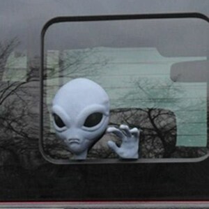 Puede incluir: Una figurita de extraterrestre gris asomándose por la ventana de un coche. El extraterrestre tiene ojos grandes y una boca pequeña. La figurita está unida a la ventana con una ventosa.