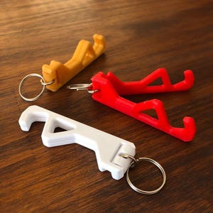 May include: Three keychains with different coloured phone stand attachments. The keychains are silver and the phone stands are yellow, red and white.