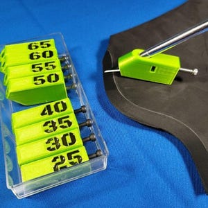 Puede incluir: Una caja de plástico transparente con bloques de plástico verde numerados del 25 al 65. Los bloques están dispuestos en dos filas. Una herramienta de plástico verde con una varilla de metal se muestra en el fondo.