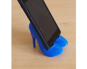 Puede incluir: Un soporte para teléfono de color azul impreso en 3D con forma de par de tacones altos. El soporte sostiene un teléfono inteligente negro.