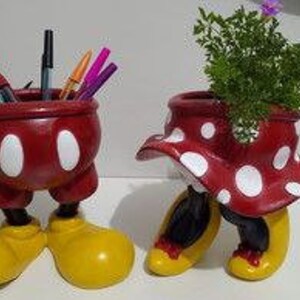 Puede incluir: Un par de macetas de cerámica con los personajes de Mickey Mouse y Minnie Mouse. La maceta de Mickey Mouse es de color rojo y blanco con lunares y tiene la forma de las piernas y los zapatos de Mickey Mouse. La maceta de Minnie Mouse es de color rojo y blanco con lunares y tiene la forma de las piernas y los zapatos de Minnie Mouse. La maceta de Minnie Mouse tiene una pequeña planta creciendo en ella.