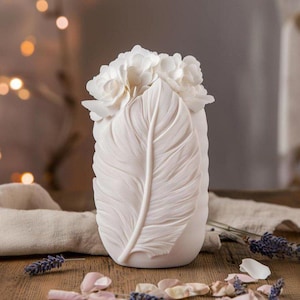 Op de afbeelding: Witte keramische vaas in de vorm van een veer met witte bloemen erop. De vaas is een decoratief woonaccessoire.
