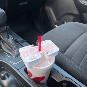May include: A white Chick-fil-A cup with a red paw print logo and a red straw. Two Chick-fil-A sauce packets are on top of the cup. The cup is in a car's cup holder.