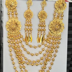 May include: A gold-tone necklace with five strands of beads and two matching earrings. The necklace features two large, ornate, circular pendants. The earrings have a smaller, circular pendant with a dangling drop.