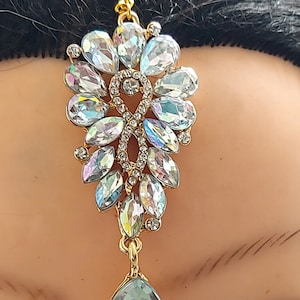 Può includere: Una catena d'oro con un ciondolo a forma di lacrima di cristallo. Il ciondolo è fatto di cristalli trasparenti con colori arcobaleno iridescenti. La catena è attaccata a un grande gruppo di cristalli trasparenti con colori arcobaleno iridescenti.