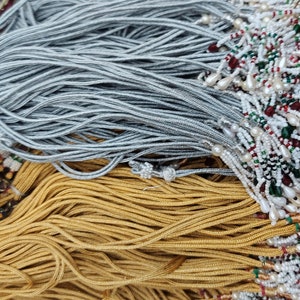 May include: A pile of silver, gold, and white beaded cords with decorative tassels. The cords are arranged in a layered pattern, with the silver cords on top, the gold cords in the middle, and the white cords on the bottom.