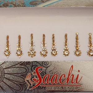May include: A pack of 10 gold and white jeweled bindi stickers. The bindi stickers are arranged in two rows of five. The pack is labelled "Saachi Bridal Bindi".
