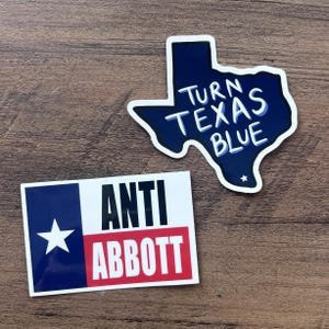 Puede incluir: Dos pegatinas sobre una superficie de madera. Una es un diseño azul en forma de Texas con las palabras "TURN TEXAS BLUE" en blanco. La otra tiene el diseño de la bandera de Texas con las palabras "ANTI ABBOTT" en negro y rojo.