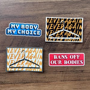 May include: Four stickers with text on a wooden surface. One sticker reads "MY BODY MY CHOICE" in blue and white. Two stickers say "NEVER AGAIN" with a hanger graphic. The last sticker says "BANS OFF OUR BODIES" in red and white.