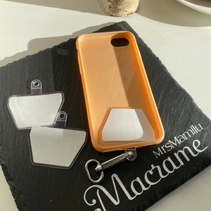 Könnte beinhalten: Eine pfirsichfarbene Handyhülle mit einem transparenten Kunststoffeinsatz und einem silbernen Metallclip. Die Hülle liegt auf einer schwarzen Schieferfläche mit dem Text "MrsMamilu Macrame" in weiß.