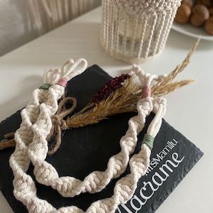 Könnte beinhalten: Zwei weiße Makramee-Schnüre mit rosa und grünen Akzenten sind auf einer schwarzen Schieferfläche aufgewickelt. Der Text "Makramee" ist in Weiß auf den Schiefer geschrieben.