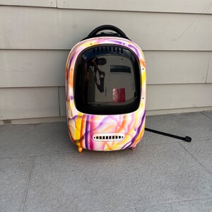 May include: A colorful, abstractly patterned pet carrier backpack with a black handle and a clear, round window. The backpack is designed to carry a small pet.