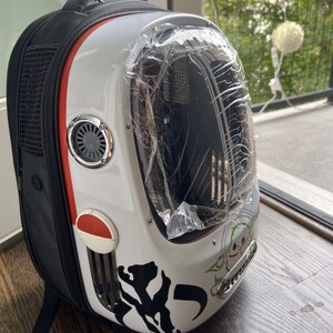 Puede incluir: Una mochila porta mascotas blanca y negra con una ventana de pl&aacute;stico transparente. La mochila tiene una franja roja y un ventilador de ventilaci&oacute;n plateado. La mochila tiene un gr&aacute;fico en blanco y negro de un personaje de Star Wars en el lateral.